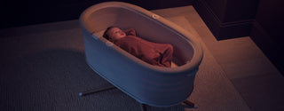 A baby sleeping in a light grey Elvie Rise smart bassinet on a stand, placed on a textured rug in a dimly lit room.