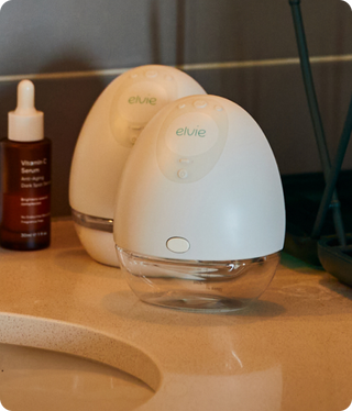 Two white elvie breast pumps on a bathroom counter with a bottle of Vitamin C serum in the background.