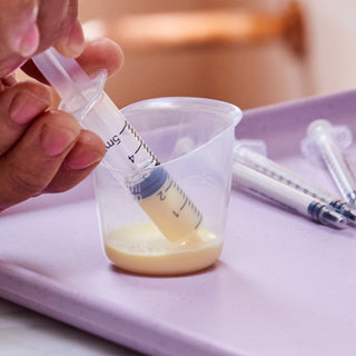 Person using a syringe to draw colostrum from a beaker on a purple surface.