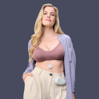 Woman wearing a bra with Elvie Stride 2 breast pump inserted and beige pants with a pump hub attached on a dark background