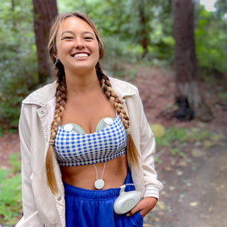 Woman in a checkered top with Elvie Stride breast pump in-bra and blue pants standing in a forest
