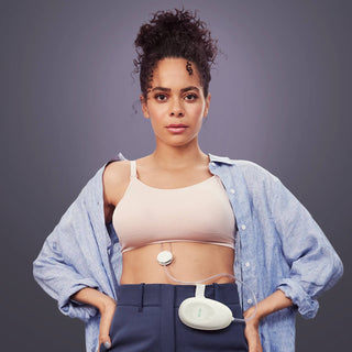 Woman wearing a Elvie Stride breast pump in-bra device with hub attached to trousers against a gray background
