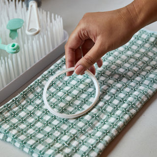 Hand holding a white Elvie Wave Gasket over a green and white checkered cloth with a baby bottle drying rack in the background.